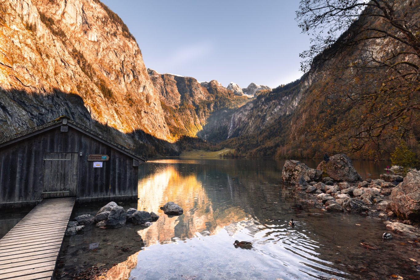 Hütte am Obersee Hölzerne Hütte am Ufer eines ruhigen Gewässers, umgeben von Bergen und herbstlichen Bäumen.